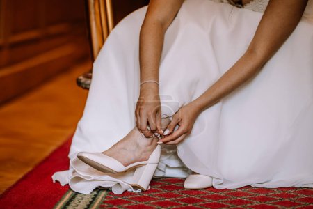 Valmiera, Latvia - August 19, 2023 - Close-up of a bride sitting down, buckling her white wedding shoe on her foot, dressed in a beautiful white gown.