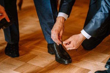 Valmiera, Letonia - 19 de agosto de 2023 - Acercamiento de un hombre atando los cordones del zapato de vestir negro de otro hombre en un pasillo de madera calurosamente iluminado.