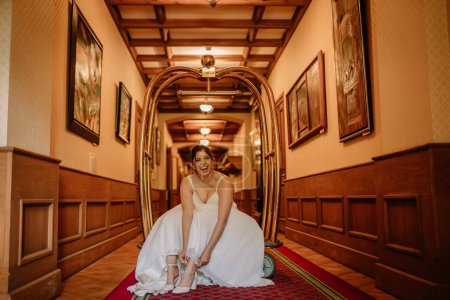 Photo for Valmiera, Latvia - August 19, 2023 - A joyful bride sits on the floor in a hotel hallway, laughing while putting on her white wedding shoes, framed by a large golden luggage cart. - Royalty Free Image