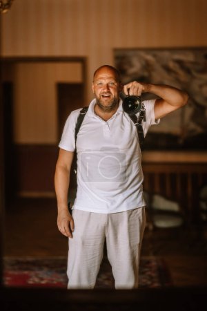 Valmiera, Latvia - August 19, 2023 - A smiling man holds a camera, ready to take a photo, wearing a white polo shirt and light pants.
