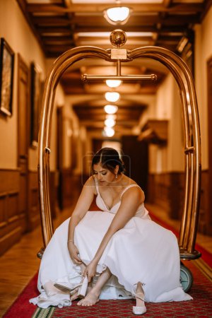 Photo for Valmiera, Latvia - August 19, 2023 - A bride in a white dress sits on a carpeted floor, putting on her wedding shoes, framed by a golden luggage cart in a hallway. - Royalty Free Image