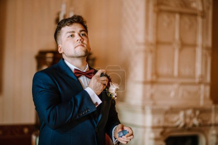Valmiera, Latvia - August 19, 2023 - A groom in a blue suit adjusts his boutonniere while holding a perfume bottle, in a decorated room.