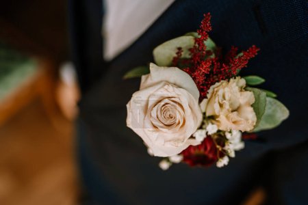 Valmiera, Latvia - August 19, 2023 - Close-up view of a groom's boutonniere on a blue suit, featuring a pale rose and red accents, with a blurred background.