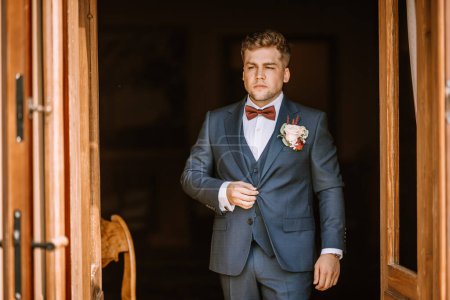 Valmiera, Lettonie - 19 août 2023 - Le marié en costume bleu se tient avec confiance dans une porte, ajustant sa veste avec une expression sérieuse.