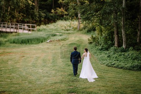 Valmiera, Latvia - August 19, 2023 - A bride and groom walk hand in hand across a grassy field towards a wooded area, with a bridge in the background.