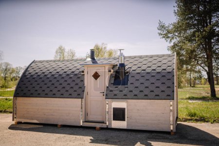 Téléchargez les photos : Une petite cabane en bois avec un toit en bardeaux, avec deux cheminées et une porte d'entrée, située dans un cadre extérieur herbeux. - en image libre de droit