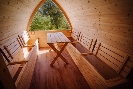 Inside a wooden cabin, a simple wooden table with benches under an arch window offering a view of lush greenery outside.