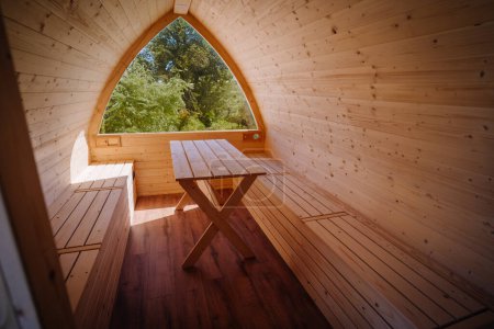 Interior view of a wooden cabin with a simple table, bench, and a large arch window framing a forest view.