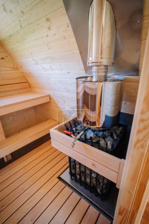 A close-up of a sauna stove with a shiny metal chimney and wooden drawer filled with stones, located inside a wooden sauna cabin with benches.