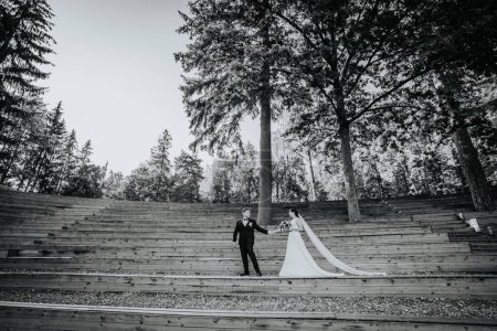 Valmiera, Lettland - 19. August 2023 - Braut und Bräutigam halten Händchen auf einem hölzernen Amphitheater im Wald, der lange Schleier der Braut hängt die Treppe hinunter.