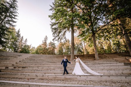 Valmiera, Lettland - 19. August 2023 - Braut und Bräutigam halten Händchen auf einem hölzernen Amphitheater im Wald, der lange Schleier der Braut hängt die Treppe hinunter.