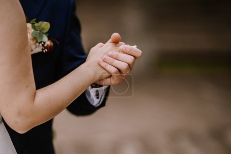 Valmiera, Latvia - August 19, 2023 - Close-up of a bride and groom holding hands, showcasing their wedding rings and the groom's boutonniere.