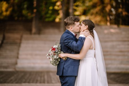 Photo for Valmiera, Latvia - August 19, 2023 - A bride and groom kiss passionately on wooden steps outdoors, holding a bouquet, with a blurred forest background. - Royalty Free Image