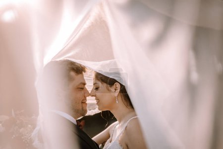 Valmiera, Lettland - 19. August 2023 - Braut und Bräutigam eng unter dem Brautschleier, beleuchtet von sanftem Gegenlicht, das eine romantische Atmosphäre schafft.