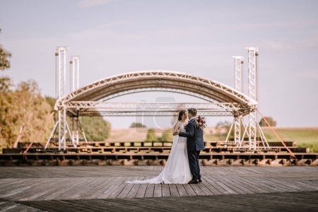 Valmiera, Latvia - August 19, 2023 - Bride and groom embrace on a wooden stage under a large metal arch in a rural setting.