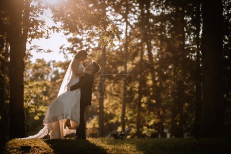Photo for Valmiera, Latvia - August 19, 2023 - Bride and groom stand among sunlit trees, embracing and kissing. Groom lifts bride, surrounded by vibrant natural scenery. - Royalty Free Image