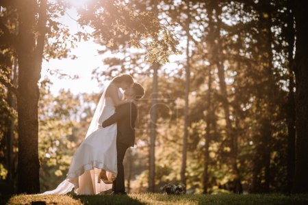Photo for Valmiera, Latvia - August 19, 2023 - Bride and groom share a kiss in a sunlit forest, with the groom lifting the bride, surrounded by trees and sunlight filtering through leaves. - Royalty Free Image