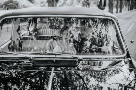 Valmiera, Lettonie - 19 août 2023 - Mariée et marié en voiture classique, vue à travers le pare-brise, expressions sérieuses, voyage, photo noir et blanc.