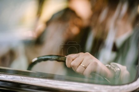 Valmiera, Latvia - August 19, 2023 - Close-up of a bride's hand on a steering wheel, wearing a wedding ring, through a car window.