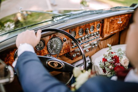 Photo for Valmiera, Latvia - August 19, 2023 - Groom driving a vintage car, hand on steering wheel, dashboard visible, wedding bouquet on seat. - Royalty Free Image