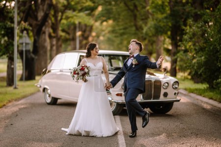 Valmiera, Latvia - August 19, 2023 - Joyful bride and groom walking hand in hand on a road, with a vintage car behind, groom jubilantly raising his arm.