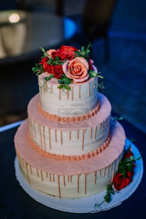 Valmiera, Letonia - 19 de agosto de 2023 - Un pastel de bodas de tres niveles adornado con rosas rojas y rosas en la parte superior, y glaseado decorativo.