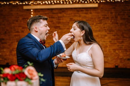 Valmiera, Lettland - 19. August 2023 - Eine Braut und ein Bräutigam füttern einander während ihres Hochzeitsempfangs glücklich mit Kuchen, im Hintergrund eine Ziegelwand und Lichter.