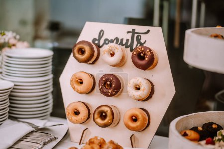 A display of assorted donuts on a wooden board, labeled "donuts," featuring various glazes and toppings, in a party setting with plates and utensils in the background.