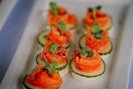 Elegant appetizers on a white dish, featuring smoked salmon rolls on cucumber slices, topped with fresh herbs and black sesame seeds.