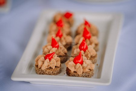 Row of pt canapes on a white plate, each topped with a vibrant red pepper garnish, served on dark bread slices.