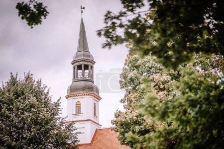 Valmiera, Latvia - August 25, 2023 - steeple of a traditional church with a weathervane on top, surrounded by green leafy trees under a cloudy sky.