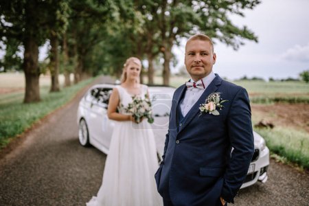 Valmiera, Latvia - August 25, 2023 - Groom in focus standing beside a luxury car with bride slightly out of focus in the background under a canopy of trees.