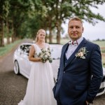 Valmiera, Latvia - August 25, 2023 - Groom in focus standing beside a luxury car with bride slightly out of focus in the background under a canopy of trees.
