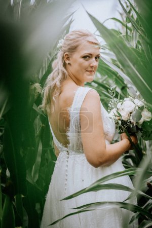Valmiera, Lettland - 25. August 2023 - Eine Braut mit einem Strauß blickt zurück, während sie inmitten großer grüner Kornhalme steht.