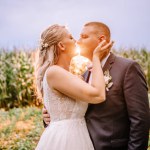 Valmiera, Latvia - August 25, 2023 - Bride and groom share a tender kiss in a sunlit field, with warm sunlight and a lush cornfield backdrop. Copy space on the right.