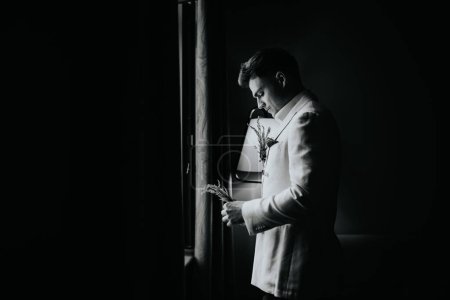 Riga, Letonia, - 26 de agosto de 2024 - Un novio con traje blanco sostiene un boutonniere, de pie cerca de una ventana en una habitación con poca luz.