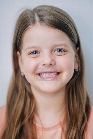 Une jeune fille souriante aux longs cheveux bruns et aux yeux bleus, portant une chemise couleur pêche et de petites boucles d'oreilles en forme de c?ur.