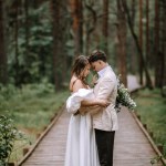 Riga, Latvia, - August 26, 2024 - Bride and groom embracing on a wooden path in a lush forest, with the bride holding a bouquet.