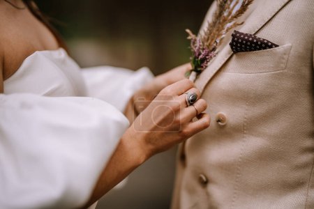 Riga, Latvia, - August 26, 2024 - Close-up of a bride adjusting a boutonniere on the groom's beige suit jacket.