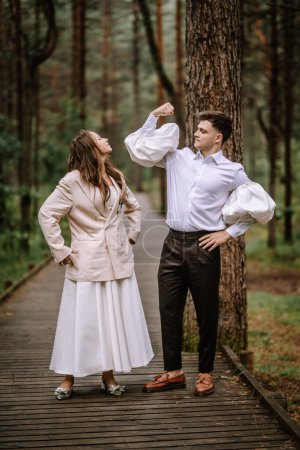 Riga, Lettland, - 26. August 2024 - Braut und Bräutigam posieren spielerisch und zeigen Muskeln auf einem Waldweg.