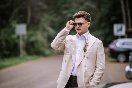 Riga, Latvia, - August 26, 2024 - Stylish man adjusting sunglasses outdoors, wearing a beige suit with a floral boutonniere.