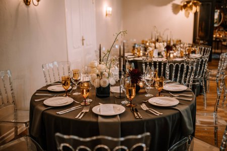 Riga, Latvia, - August 26, 2024 - Elegant indoor wedding reception setting with black tablecloths, white and dark floral centerpieces, gold-rimmed glasses, and clear chairs.