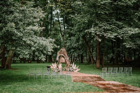 Riga, Lettland, - 26. August 2024 - Hochzeitszeremonie im Freien in einem Wald mit einem natürlichen Bogen aus getrockneten Palmen und Blumen, klaren Stühlen und einem rustikalen Teppich auf dem Rasen.