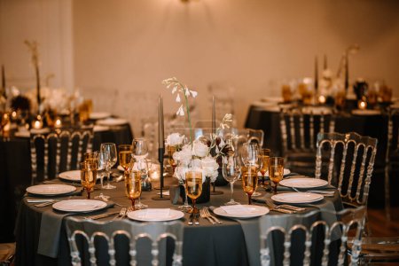 Riga, Latvia, - August 26, 2024 - Elegant wedding reception table setting with black tablecloths, golden glassware, floral centerpieces, and clear chiavari chairs in a warmly lit room.