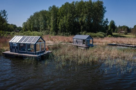 Two quaint floating houses rest on a serene lake surrounded by lush vegetation under a clear blue sky.