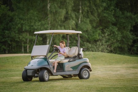 A young girl sits in a golf cart on a grassy field, smiling and holding the steering wheel, with a background of lush green trees.