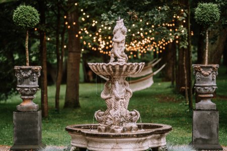 Riga, Latvia, - August 26, 2023 - Ornate stone fountain with fish sculptures in a lush garden, surrounded by trees, string lights, and topiary, creating a serene and enchanting atmosphere.