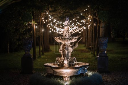 Riga, Latvia, - August 26, 2023 - An illuminated stone fountain in a garden at night, surrounded by trees and string lights, creating a serene and magical atmosphere.