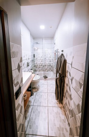 Blome, Lettland - 11. September 2023 - Ein modernes Badezimmer mit geometrisch gemusterten Fliesen, einer begehbaren Dusche, einem weißen Waschbecken, einer Toilette und zwei Bademänteln, die an der Wand hängen.