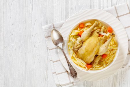 hot chicken soup of whole chicken, egg noodles, celery, onion and carrot in white bowl on wood table with napkin and spoon, flat lay, free space